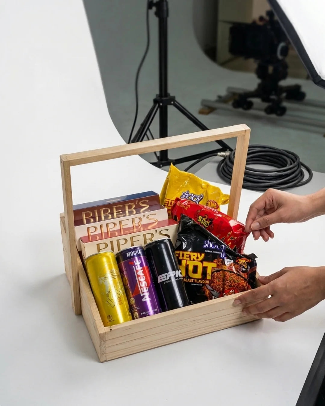 Wooden basket with snacks and drinks on a white surface, with a person's hand reaching in.